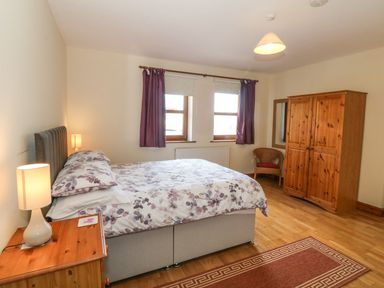 A bedroom with a bed bedside table lamp two windows purple curtains wooden wardrobe and a chair at Bwthyn Clyd in Llanddaniel Fab