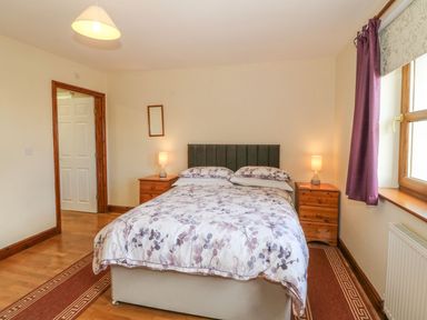 A bedroom with a double bed two bedside tables with lamps a window with purple curtains and a door at Bwthyn Clyd in Llanddaniel Fab