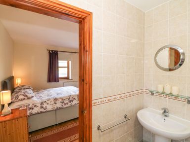 A bedroom with a bed and nightstand visible through a wooden door next to a bathroom sink with a round mirror at Bwthyn Clyd in Llanddaniel Fab
