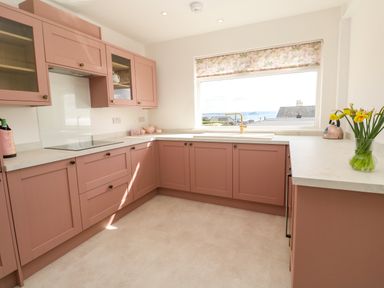 A kitchen with pink cabinets white countertops a window with a roller blind and a vase of yellow flowers at Karenza in Penzance