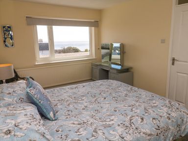 A bedroom with a bed covered in floral bedding a window showing an outside view a lamp a dressing table with mirrors and a closed door at Karenza in Penzance