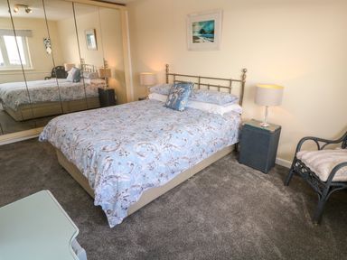 A bedroom with a bed with floral bedding beside a nightstand with a lamp and a chair with a cushion at Karenza in Penzance