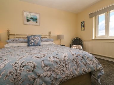 A bedroom with a bed covered in blue floral bedding a lamp on a side table a window and a chair at Karenza in Penzance