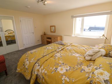 A bedroom with a floral yellow bedspread a wooden dresser with a mirror and a window showing rooftops at Karenza in Penzance