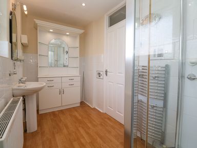 A bathroom with a white sink and cabinet a mirror a white door wooden floor and a glass shower door at Karenza in Penzance