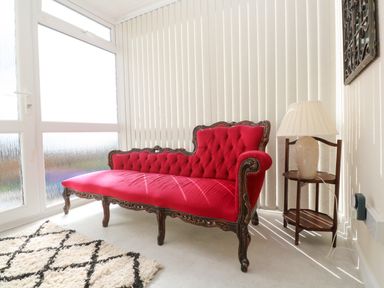 A room with a red upholstered wooden sofa a small wooden side table with a lamp and vertical blinds covering the windows at Karenza in Penzance
