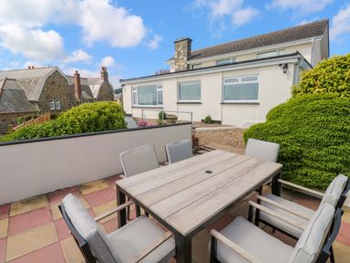 An outdoor patio area with a wooden table and six cushioned chairs near a white house with bushy greenery at Karenza in Penzance