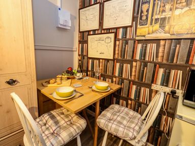 A small dining area with a wooden table set for two with bowls and cutlery two cushioned chairs and framed maps on a wallpapered wall with book spines at The Cabin at Little Timbers in Ringwood
