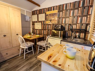 A small kitchen with a wooden table set for two a sink microwave and a fridge with wallpaper showing a bookshelf at The Cabin at Little Timbers Ringwood