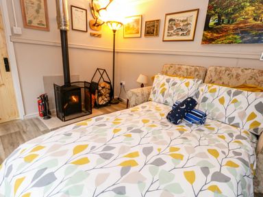 A bedroom with patterned bedspread a wood stove firewood and framed pictures on the wall at The Cabin at Little Timbers in Ringwood