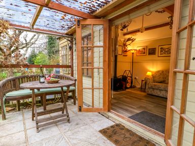 A patio with a wooden table and bench outside an open door showing a living room with a sofa and lamp at The Cabin at Little Timbers in Ringwood
