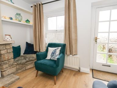 A corner of a room with a teal armchair with a cushion a stone seating area with cushions shelves with glass vases beige curtains a window and a glass door at Honeypot Cottage in Swanage