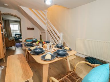 A dining area with a wooden table set for six under stairs next to a wicker chair and a radiator at Honeypot Cottage in Swanage