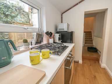A kitchen counter with a gas stove three yellow mugs a cutting board and a teal kettle next to a window at Honeypot Cottage in Swanage