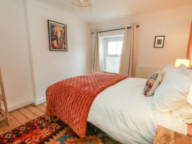 A bedroom with a double bed with white bedding and an orange quilt with a window with cream curtains at Honeypot Cottage in Swanage