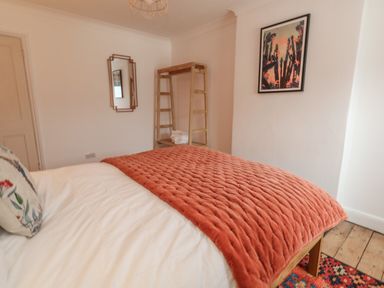 A bedroom with a bed covered with a white duvet and an orange blanket next to a wooden shelf with folded towels and a framed picture on the wall at Honeypot Cottage in Swanage