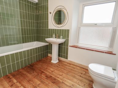 A bathroom with green tiled walls a bathtub a pedestal sink a circular mirror a toilet and a window at Honeypot Cottage in Swanage