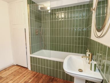 A bathroom with green tiled walls a bathtub with a glass shower screen a white sink with a faucet and a round mirror above it at Honeypot Cottage in Swanage
