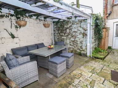 An outdoor seating area with a wicker table and cushions under a pergola with hanging baskets at Honeypot Cottage in Swanage