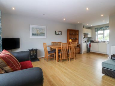 An open living area with a dining table and six chairs a small stove and a television next to a kitchen with white cabinets at Ty Nant in Abersoch