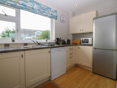 A kitchen with cream cabinets a dishwasher a stainless steel fridge a microwave toaster kettle and a window with a blue sailboat curtain at Ty Nant in Abersoch