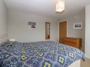 A bedroom with a patterned bedspread a wooden dresser a wooden door a rectangular mirror and two framed pictures at Ty Nant in Abersoch