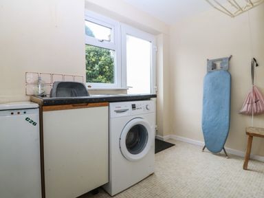 A laundry room with a washing machine a sink an ironing board a wooden stool and a door with a window at Ty Nant in Abersoch