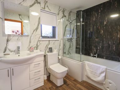 A bathroom with a bathtub shower combo toilet sink and mirror with marble patterned walls and wood flooring at Tealby in Horning
