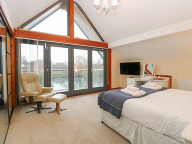 A bedroom with a bed a chair a dressing table and large windows overlooking water at Tealby in Horning
