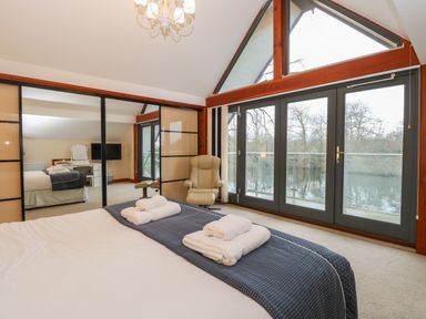 A bedroom with a bed, folded towels, an armchair, large windows overlooking trees and water, and mirrored sliding closet doors at Tealby in Horning