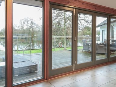 Glass doors with wooden frames overlooking a wooden deck with outdoor seating and a railing by a body of water at Tealby in Horning
