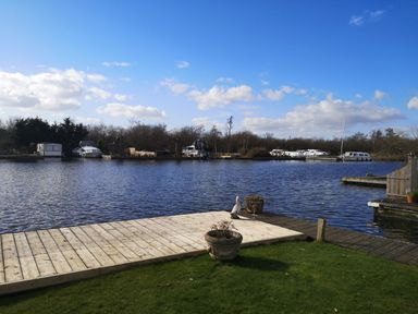 A view of a river with boats docked and a wooden deck with pots on a grassy area at White Gables in Horning