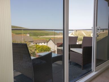 A balcony with wicker chairs and a glass table overlooking rooftops and hills at Atlantic View in Holywell Bay