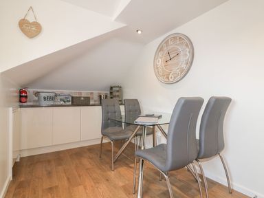 A dining area with a glass table and four gray chairs under a slanted ceiling with a wall clock and a countertop with storage cabinets