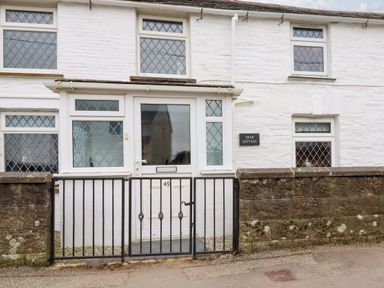 The exterior of a white stone cottage with diamond-patterned windows and a black metal gate at Crab Cottage in Delabole