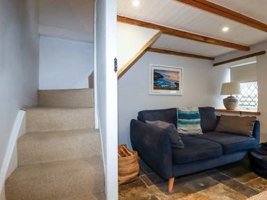 A room with carpeted stairs a blue sofa with cushions a lamp on the windowsill and a framed picture on the wall at Crab Cottage in Delabole