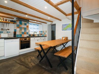 A kitchen with white cabinets a black countertop wooden table with benches and carpeted stairs at Crab Cottage in Delabole