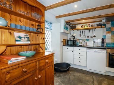 A kitchen with wooden shelves holding mugs and glasses and white cabinets with kitchen appliances at Crab Cottage in Delabole