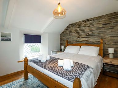 A bedroom with a wooden bed frame and white bedding with folded towels on top and a stone accent wall at Crab Cottage in Delabole