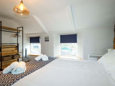 A bedroom with a bed with rolled towels two windows with blue blinds and a shelving unit at Crab Cottage in Delabole