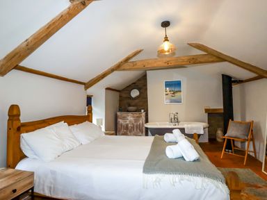 A bedroom with a wooden bed frame white bedding rolled towels on bed a bathtub a wooden chair and exposed ceiling beams at Crab Cottage in Delabole