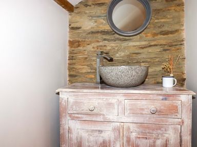 A bathroom sink with a round mirror on a stone wall above a wooden cabinet with drawers at Crab Cottage in Delabole