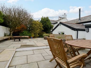 An outdoor patio with wooden chairs and table stone flooring and surrounding bushes at Crab Cottage in Delabole