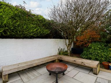 An outdoor patio area with a wooden bench surrounding a metal fire pit with trees and shrubs in the background at Crab Cottage in Delabole
