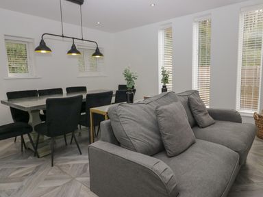 A room with a gray sofa black dining chairs a dining table with three pendant lights and windows with blinds at Edgemere Cottage in Cartmel