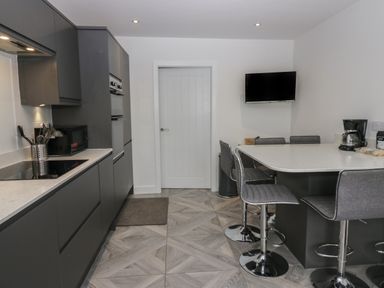 A kitchen with dark cabinets a countertop with a microwave a coffee maker chairs around a peninsula and a wall-mounted television at Edgemere Cottage in Cartmel