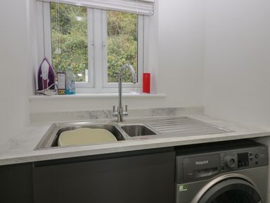 A utility room with a sink a washing machine and cleaning items on the windowsill at Edgemere Cottage in Cartmel