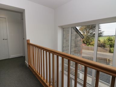 A hallway with a wooden railing and large windows overlooking a street and trees at Edgemere Cottage in Cartmel