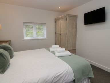 A bedroom with a bed with green pillows and a blanket a wardrobe a window with blinds and a wall-mounted television at Edgemere Cottage in Cartmel