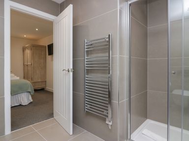 A bathroom with a wall-mounted towel rack and a glass shower next to a door leading to a bedroom with a wardrobe and a wall-mounted television at Edgemere Cottage in Cartmel